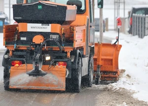 Ein orangefarbenes Winterdienst-Fahrzeug räumt Schnee und streut Salz auf einer verschneiten Straße, um die Sicherheit bei Winterwetter zu gewährleisten.
