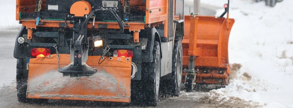 Ein orangefarbenes Winterdienst-Fahrzeug räumt Schnee und streut Salz auf einer verschneiten Straße, um die Sicherheit bei Winterwetter zu gewährleisten.