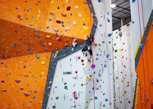Eine Person klettert an einer bunt markierten Kletterwand in einer Kletterhalle. Die Wandfläche ist in Grau und Orange gestaltet und mit farbigen Klettergriffen in Blau, Rot, Gelb, Lila, Grün und Weiß versehen. Die Person trägt Kletterschuhe, ein Klettergeschirr und ist durch ein Seil gesichert. Rundherum sind weitere Routen mit unterschiedlich farbigen Griffen sichtbar.