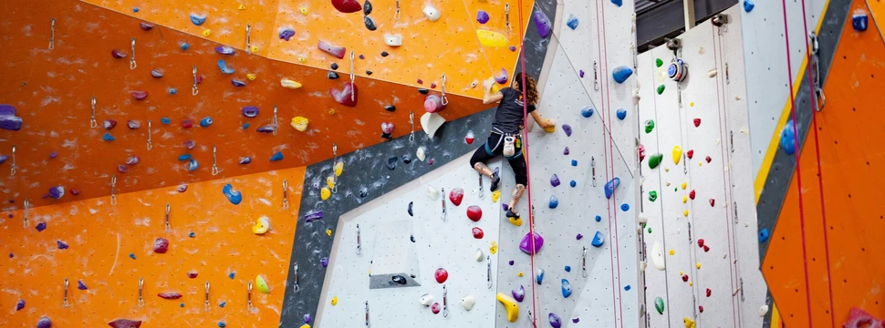 Eine Person klettert an einer bunt markierten Kletterwand in einer Kletterhalle. Die Wandfläche ist in Grau und Orange gestaltet und mit farbigen Klettergriffen in Blau, Rot, Gelb, Lila, Grün und Weiß versehen. Die Person trägt Kletterschuhe, ein Klettergeschirr und ist durch ein Seil gesichert. Rundherum sind weitere Routen mit unterschiedlich farbigen Griffen sichtbar.