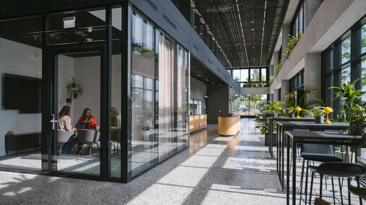 Galerie-Bild 1: Modernes Bürogebäude mit Glaswänden, grünen Pflanzen und Menschen in Besprechung bei Tageslicht.