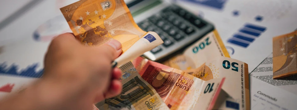 Mehrere Gelscheine verschiedenen Wertes in einer Hand mit einem Taschenrechner im Hintergrund