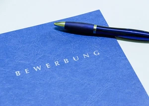 Mit einer guten Bewerbung punktet man bei Recruitern. Blaue Bewerbungsmappe, auf der ein blauer Kugelschreiber liegt
