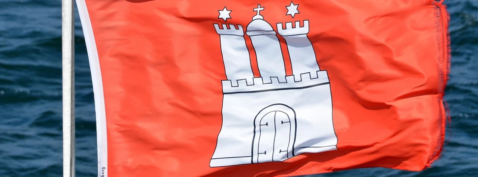 Rote Flagge mit dem weißen Hamburger Wappen (Burg mit drei Türmen und Sternen) weht im Wind vor blauem Wasser.