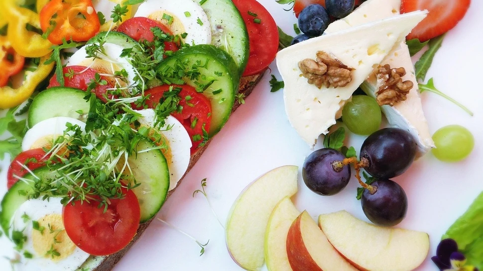 Belegtes Brot mit Gemüse und Ei, dazu Käse, Nüsse und frisches Obst auf einem Teller.