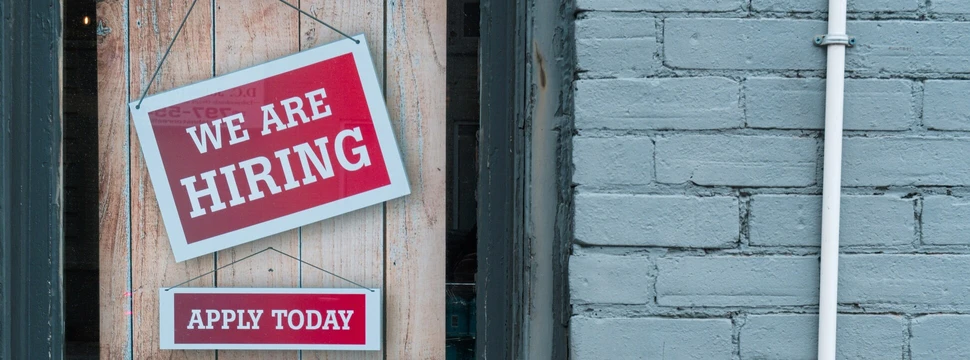 Ein Schaufenster mit roten Schildern, die mit weißer Schrift den Text "We are hiring" und "Apply today" zeigen.