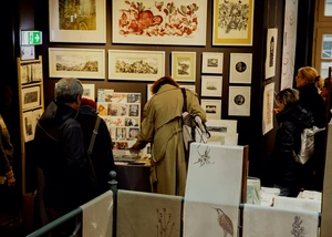 Besucher betrachten kunstvolle Drucke und handgefertigte Werke in einem stimmungsvoll beleuchteten Ausstellungsraum mit gerahmten Bildern an den Wänden.