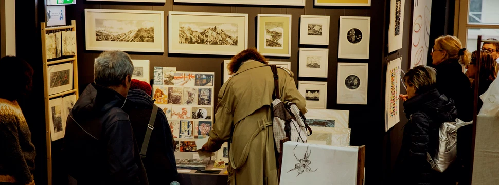 Besucher betrachten kunstvolle Drucke und handgefertigte Werke in einem stimmungsvoll beleuchteten Ausstellungsraum mit gerahmten Bildern an den Wänden.