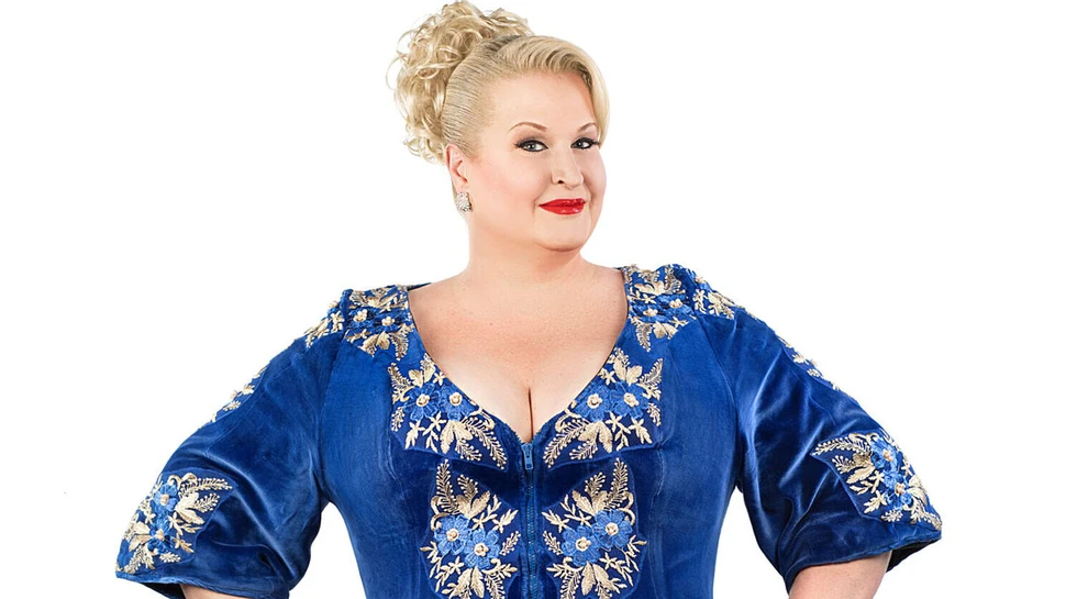 Daphne de Luxe Eine Frau zwischen 50 und 60 in einem blauen Kleid mit Blumenstickereien vor einem weißen Hintergrund