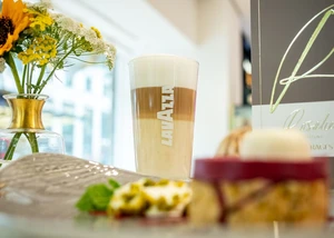 Latte Macchiato in einem LAVAZZA Glas, davor unscharf ein Stück Kuchen