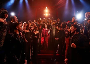 Szenenbild aus dem Musical mit einer Frau im roten Kleid und anderen festlich gekleideten Menschen