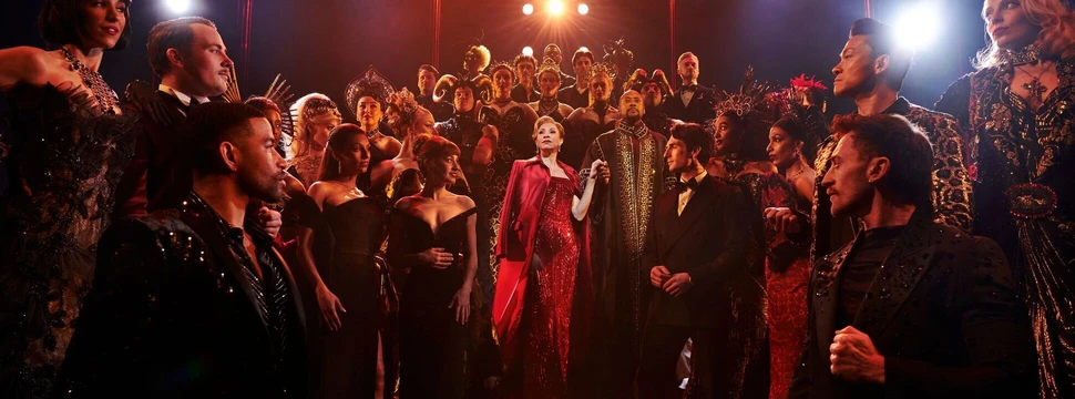 Szenenbild aus dem Musical mit einer Frau im roten Kleid und anderen festlich gekleideten Menschen