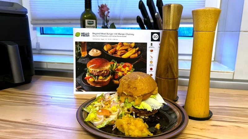 dunkler Teller mit Beyond Meat Burger und Mango-Chutney, daneben Pfeffermühlen und ein Messerblock