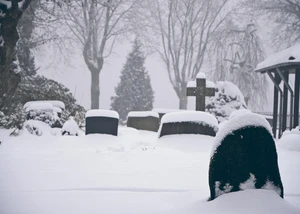 Schneebedeckter Friedhof mit Grabsteinen und einem Kreuz, umgeben von kahlen Bäumen in einer winterlichen, nebligen Atmosphäre.