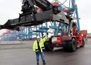 Ein Mann mit Warnjacke vor einem Kran, der ein Geschenk im Greifarm hält