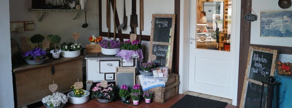 Der Eingangsbereich des Hofladens Bimöhlen mit Blumen, Deko und rustikalem Charme lädt ein. Durch die Tür sind frische Produkte sichtbar.