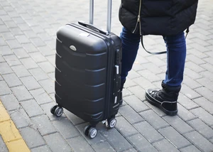 Eine Person mit schwarzer Jacke und Jeans steht auf einem gepflasterten Boden, neben ihr ein schwarzer Rollkoffer.