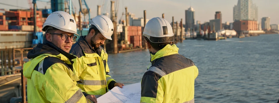 rei Bauarbeiter in Sicherheitskleidung studieren Baupläne am Containerhafen mit Kränen und Stadtsilhouette.