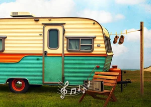 Aber bitte mit Schlager - Komödie im hoftheater Hamburg Ein alter Campingwagen steht auf einem Wiesenabschnitt am Strand, davor steht ein Stuhl mit einem alten Radio darauf und nebendran eine Wäscheleine mit einem Paar Sandalen, Links im Bild ist ein Schriftzug mit dem Titel des Theaterstücks.