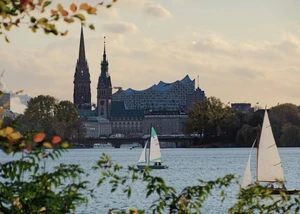 Segelboote auf der Hamburger Außenalster, dahinter die Skyline mit Kirchtürmen und der Elbphilharmonie im Abendlicht.