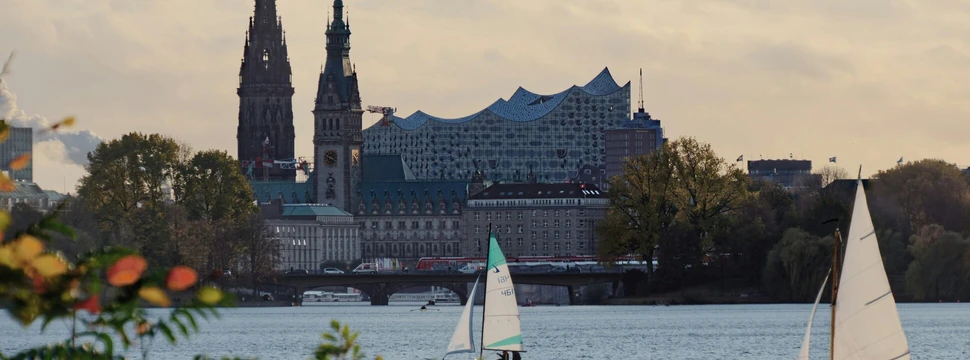 Segelboote auf der Hamburger Außenalster, dahinter die Skyline mit Kirchtürmen und der Elbphilharmonie im Abendlicht.