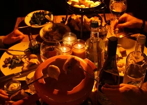 Gemütlich gedeckter Tisch im Halbdunklen mit Käsefondue-Topf, Kerzen und Flaschen