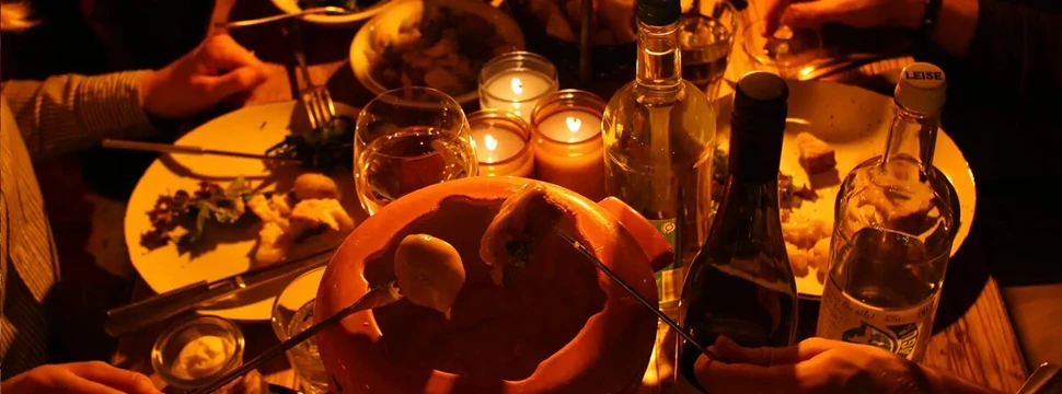Gemütlich gedeckter Tisch im Halbdunklen mit Käsefondue-Topf, Kerzen und Flaschen