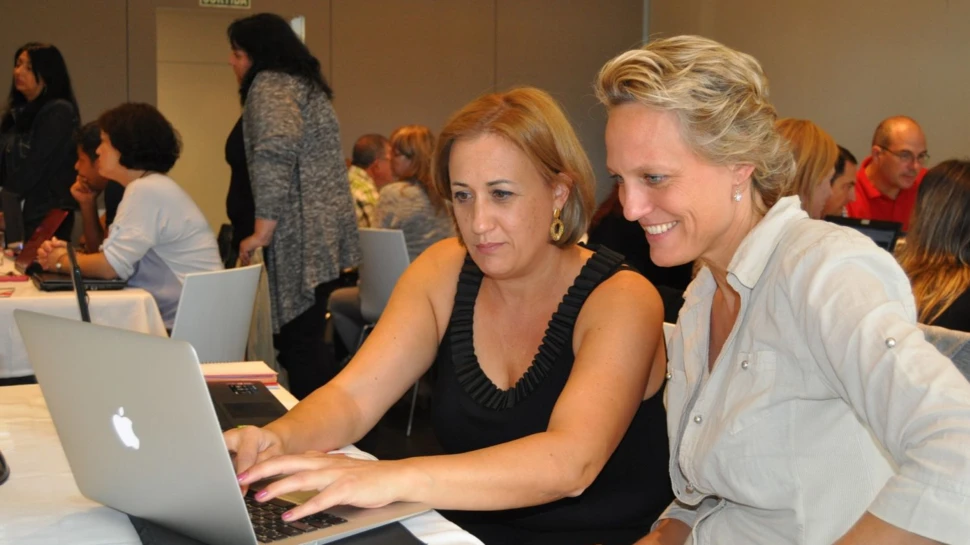 zwei Frauen sitzen vor einem Laptop im Raum mit weiteren Menschen