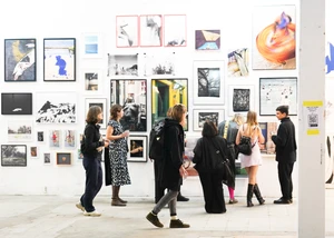 Mehrere Personen betrachten in einer Galerie zahlreiche Fotografien und Kunstwerke, die dicht an einer weißen Wand angebracht sind. Die Ausstellung zeigt eine vielfältige Mischung aus Schwarz-Weiß- und Farbfotografien sowie abstrakter Kunst. Einige Besucher stehen in kleinen Gruppen zusammen und unterhalten sich, andere schauen aufmerksam einzelne Bilder an. Die Atmosphäre wirkt lebendig und interessiert.