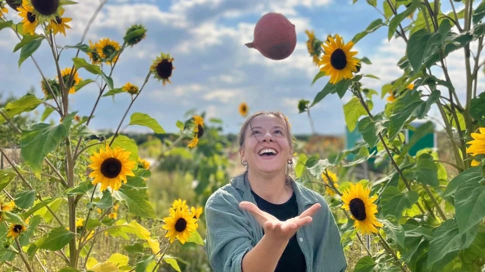 Frau jongliert mit einem Kürbis, neben ihr wachsen Sonnenblumen