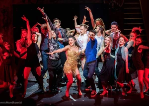 Energiegeladene Musical-Aufführung mit tanzendem Ensemble in dramatischem roten Bühnenlicht Musical-Ensemble tanzt energisch auf der Bühne in rotem Licht, Hauptdarstellerin im goldenen Kleid im Zentrum