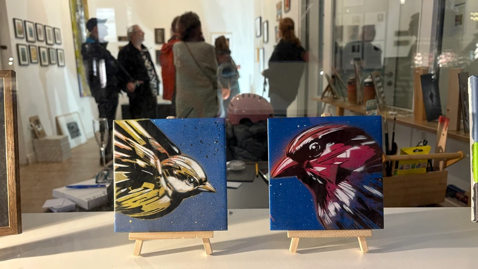 Im Vordergrund zwei quadratische Kunstwerke mit Vogelmotiven, im Hintergrund Besucher der Ausstellung