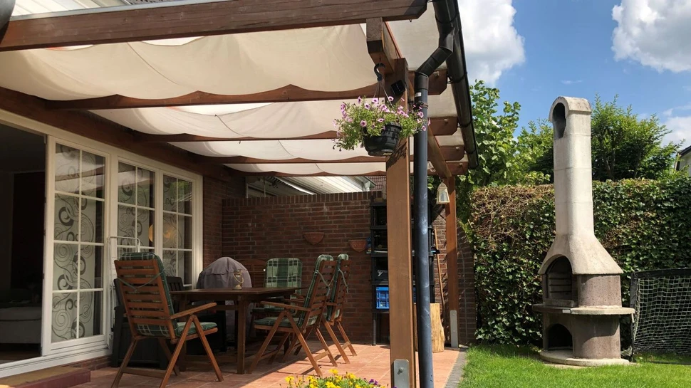 Terrassenüberdachung aus Holz mit hellen Sonnensegeln und Gartenmöbeln darunter