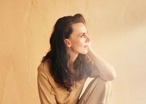 Anna Depenbusch in beigem Outfit sitzt nachdenklich vor hellem Hintergrund.