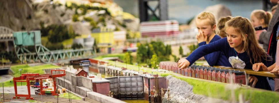 Kinder betrachten fasziniert eine detaillierte Modelleisenbahnanlage mit Zügen, Gebäuden und grüner Landschaft.