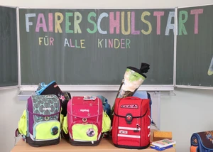Drei Schulranzen stehen auf einem Tisch vor einer Tafel mit dem Schriftzug "Fairer Schulstart für alle Kinder"