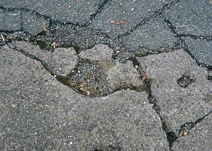 Eine Nahaufnahme einer beschädigten Asphaltstraße mit sichtbaren Rissen und Schlaglöchern, die durch Frost und Wetterbedingungen entstanden sind.