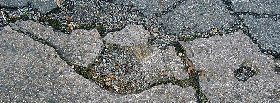 Eine Nahaufnahme einer beschädigten Asphaltstraße mit sichtbaren Rissen und Schlaglöchern, die durch Frost und Wetterbedingungen entstanden sind.