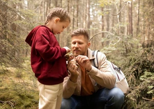Ein Mädchen ist mit mit ihrem Vater im Wald und begutachtet einen Tannenzapfen.