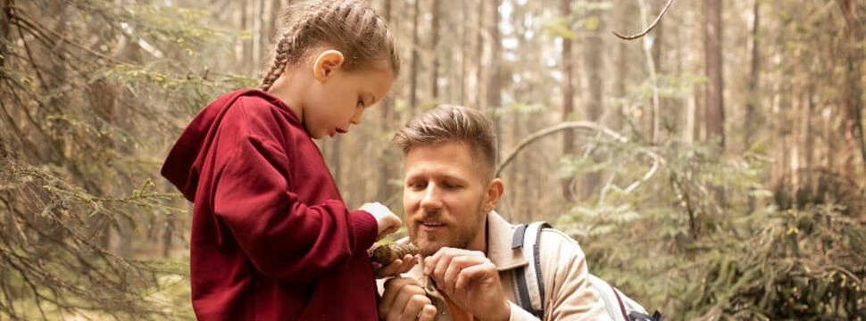 Ein Mädchen ist mit mit ihrem Vater im Wald und begutachtet einen Tannenzapfen.