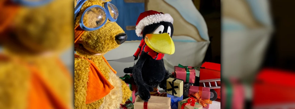 Der kleine Rabe Socke mit einer Weihnachtsmütze auf dem Kopf sitzt hinter einem kleinen Geschenkeberg