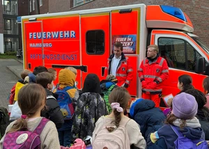Ein paar Kinder im Grundschulalter stehen vor einem Krankenwagen mit zwei Mitarbeiter des AKK