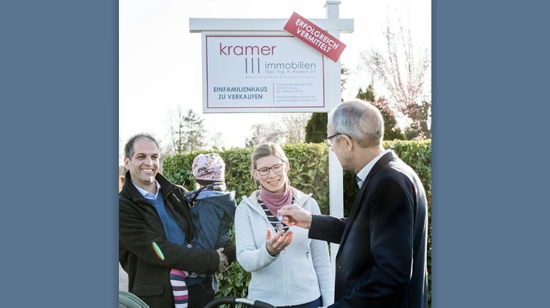 Eine Familie steht mit ihrem Makler beim Kauf des Hauses vor einem Galgen wo drauf steht: erfolgreich vermittelt.