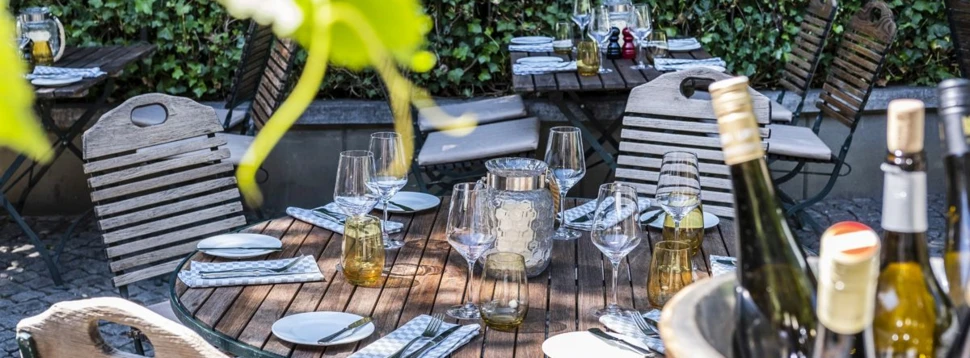 Eine stilvoll gedeckte Gartenterrasse im Weingarten Kleines Jacob lädt zum entspannten Dining im Freien ein Gedeckte Gartenterrasse mit Holztischen, Weingläsern und Weinkühler vor grüner Efeu-Wand.
