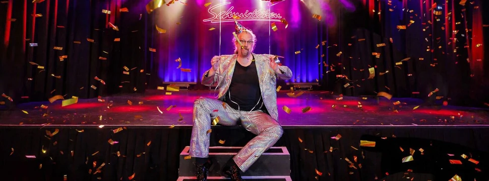 10 Jahre Schmidtchen – Geburtstagssause mit Comedy-Show und Party Mann im Glitzeranzug sitzt auf einer Bühne mit Konfetti