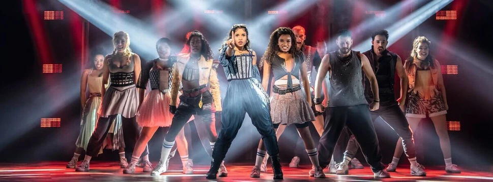 Musical-Ensemble auf Bühne mit spektakulärer Lichtshow, kreuzende Scheinwerfer und rote Beleuchtung.