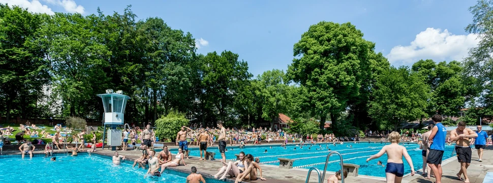 Zwei Becken im Freibad mit vielen verschiedenen Menschen und Bäumen und blauer, leicht bewölkter Himmel im Hintergrund