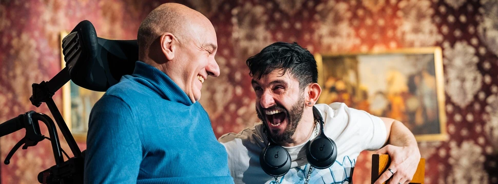 ein Mann im Rollstuhl, daneben ein lachender, jüngerer Mann
