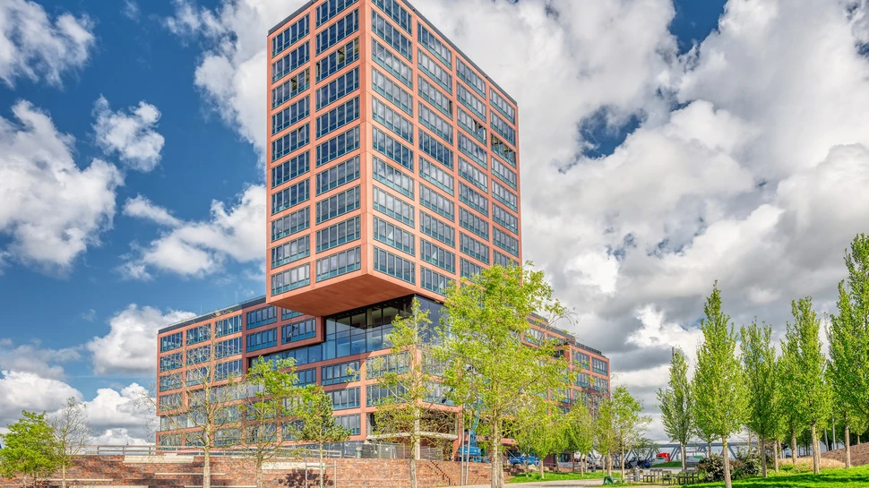 Modernes, terrakottafarbenes Bürogebäude mit auskragendem Obergeschoss in einem Park mit jungen Bäumen unter blauem Himmel.