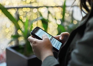 Eine Person an einem Handy in der Hand, die gerade auf einer Bank vor Pflanzen sitzt